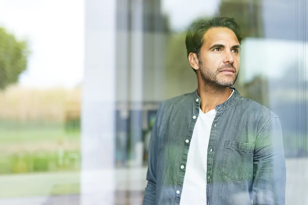 A man is standing at a window looking outside. 