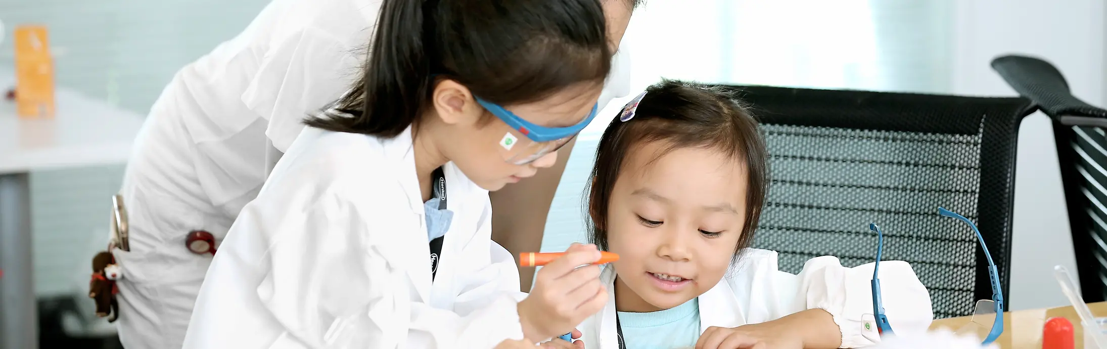 Due bambine e una donna con camici da laboratorio colorano un disegno su un tavolo